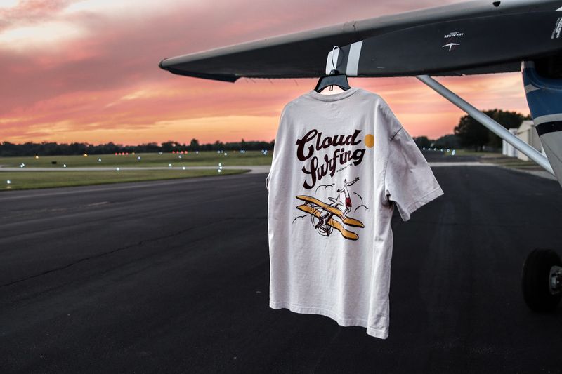 A Cloud Surfing tee hangs under a plane wing against a pink sky