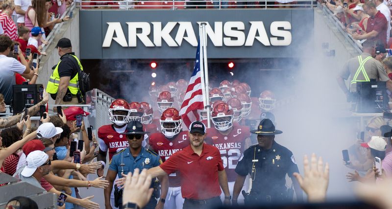Arkansas football players run out of the tunnel through smoke behind an American flag